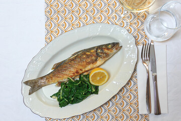 Grilled fish Seabass with spinach garnish served with a knife and fork with a glass of water and wine