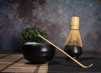 matcha tea black bowl with gray stains gray whisk and bamboo spoon on table with cane tablecloth with old green tea old green tea cement background with stains foreground photo of diner view with spac