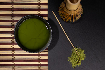 bowl of black matcha tea with gray stains whisk and bamboo spoon on table with cane tablecloth with green tea old cement background with stains close-up top view photo with space for text 