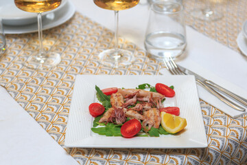 square plate with mini calamari and garnish of green leaves and cherry tomatoes served with a knife and fork with a glass of water and wine