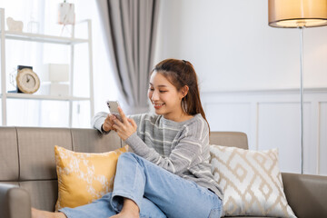 Happy and cheerful woman is using a phone sitting on a sofa.Asian young woman looking mobile phone...