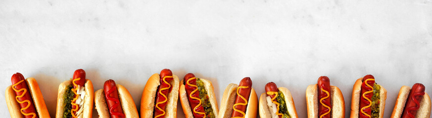 Assortment of hot dogs with a variety of toppings. Top view bottom border on a white marble background.
