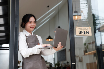 Young adorable Asian barista holding laptop while standing next to the open sign in front of her...