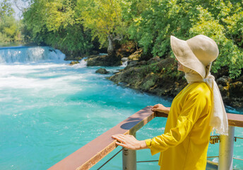 Female tourist in a yellow dress and sunhat looks at a beautiful waterfall in Manavgat, Turkey. Travel, tourism, ecology, nature, human interaction with nature.