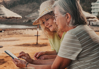 Caucasian couple of women standing at the beach looking together at smartphone. Senior and middle...
