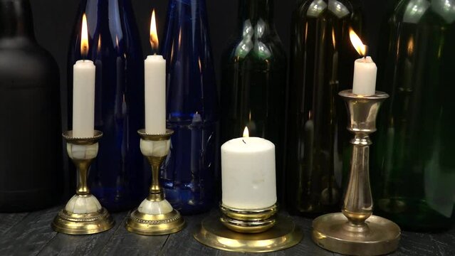 The flame of burning candles in two brass candlesticks is extinguished by a breath of wind. Burning white candles in old vintage brass and bronze candlesticks against the background of glass multicolo