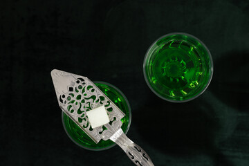 Two glasses of absinthe with a silver spoon and a cube of sugar on a green background, top view