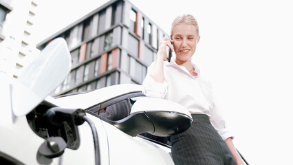 Progressive businesswoman leaning on electric car and charging station before driving around city...