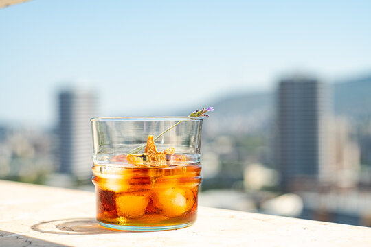 Glass of whiskey with ice and orange peel on white surface against blurred city