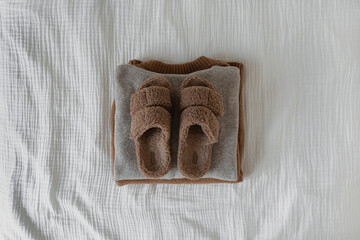 Cozy brown slippers on stack of warm wool pullovers and sweaters on white muslin cloth. Warm home wearing. Flat lay, top view