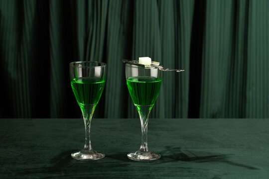 Two glasses of absinthe with a silver spoon and a cube of sugar on a green background
