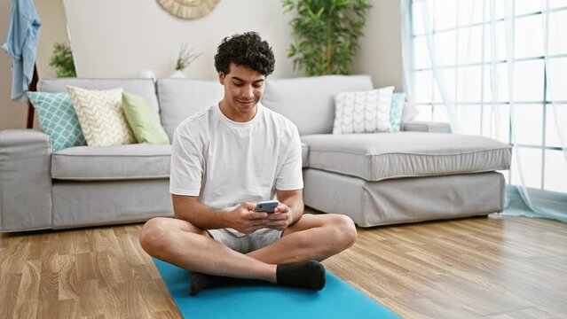 Young latin man using smartphone sitting on yoga mat at home