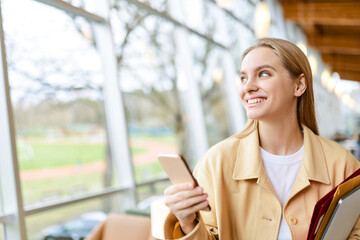 Happy girl student using mobile phone looking away walking indoors. Young smiling blonde woman holding cellphone modern tech standing in university or office advertising apps and services. Copy space