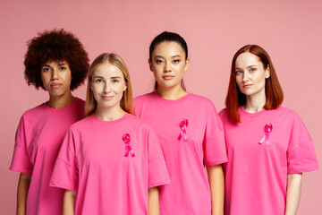 Group of serious multiracial women  wearing  t shirts with breast cancer pink ribbon isolated on pink background. Health care, support, prevention. Breast cancer awareness month concept 