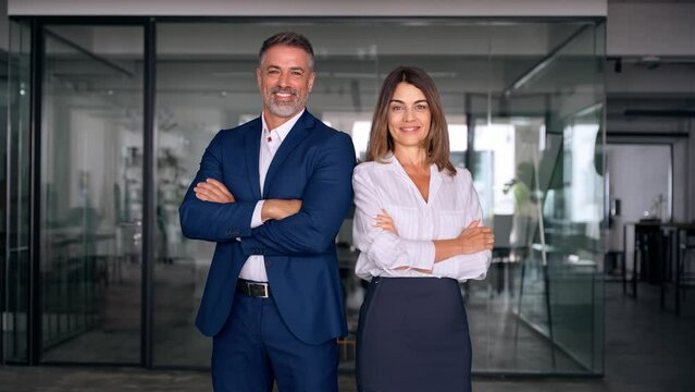 Portrait of smiling mature Latin or Indian business man and European business woman standing arms crossed in office. Two diverse colleagues, group team of confident professional business people.