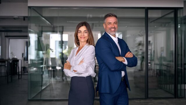 Portrait of smiling mature Latin or Indian business man and European business woman standing arms crossed in office. Two diverse colleagues, group team of confident professional business people.