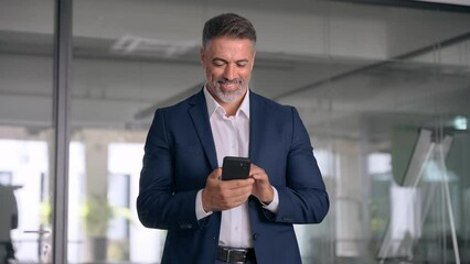 Smiling mature Latin or Indian businessman holding smartphone in office. Middle aged manager using cell phone mobile app. Digital technology application and solutions for business success development.