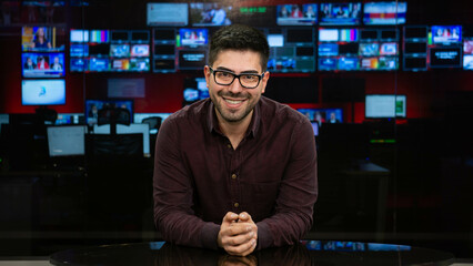 man with glasses in television studio smiling looking at camera. Television presenter, news anchor middle aged man in front of camera for TV show. © AlpEren
