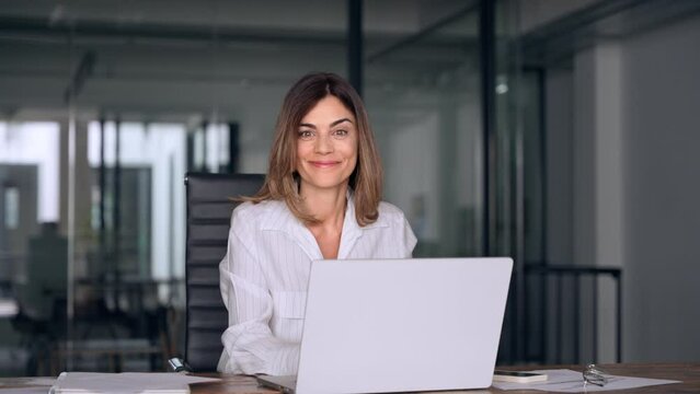 40s mid age European business woman CEO using laptop for work sitting at table in office and looking at camera. Smiling Latin Hispanic mature adult professional businesswoman using pc digital computer