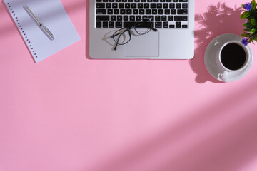 Home office workspace with a cup of coffee, a laptop, a pair of glasses, a pen, a bundle of binder paper, an artificial plant and shadows on pink background
