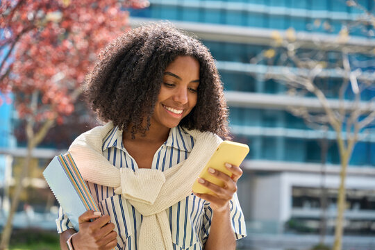 Smiling African American Girl Walking In University Park Using Mobile Cell Phone Shopping Online Or Texting Messages On Cellphone. Happy Female Student Holding Smartphone Standing Outdoors.