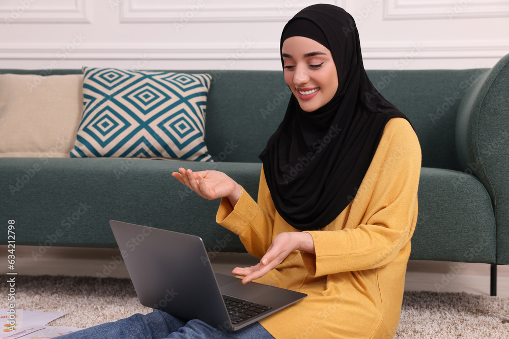 Poster Muslim woman in hijab using video chat on laptop near sofa indoors