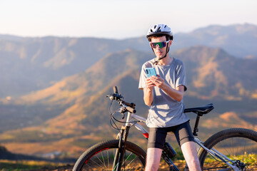 Cyclist standing with his mountain bike on the trail with mountains in the background accessing the internet with 5G or using a cyclist app on his smartphone.