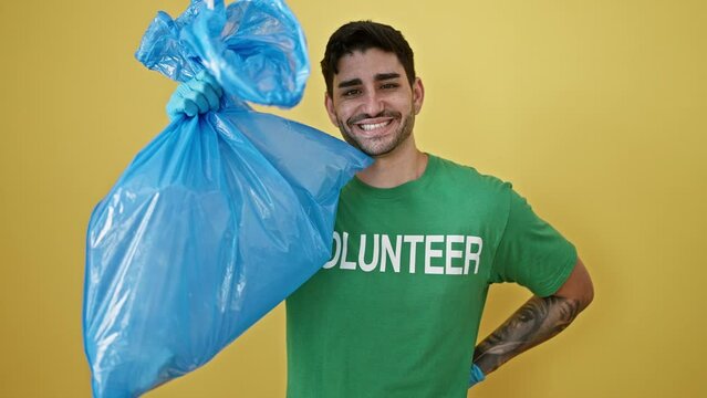 Young hispanic man volunteer wearing gloves holding garbage bag over isolated yellow background