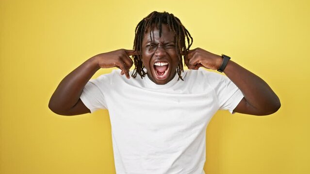 African american man screaming loudly covering ears with fingers over isolated yellow background