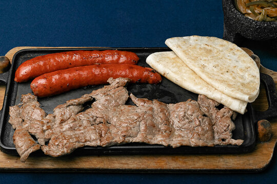 dish of Mexican roast beef and pork, accompanied by two quesadillas and molcajete sauce, on a wooden board and a matal comal on a blue table.