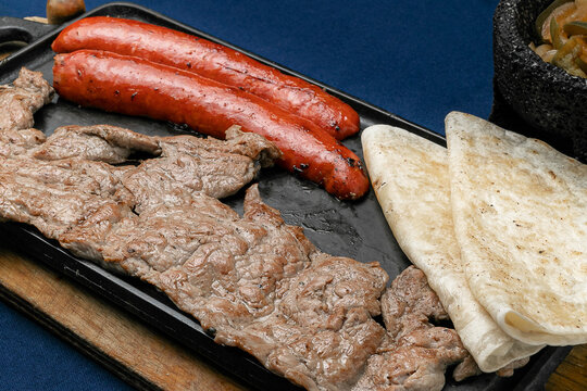 dish of Mexican roast beef and pork, accompanied by two quesadillas and molcajete sauce, on a wooden board and a matal comal on a blue table.