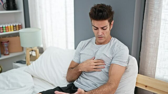 Young hispanic man sitting on bed suffering heart attack at bedroom