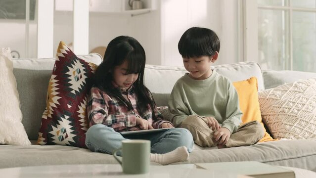 two cute asian children brother and sister sitting on family couch playing computer game using digital tablet