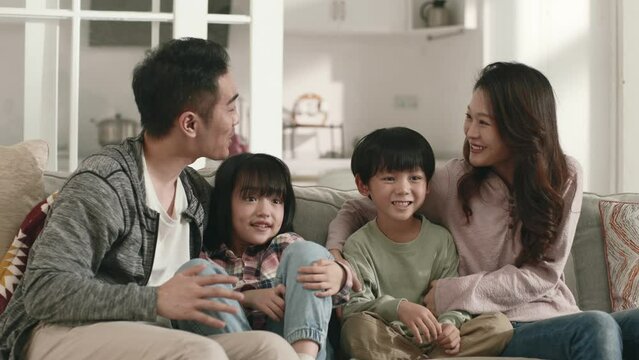 Happy Young Asian Family With Two Children Sitting On Couch At Home Watching Tv Together