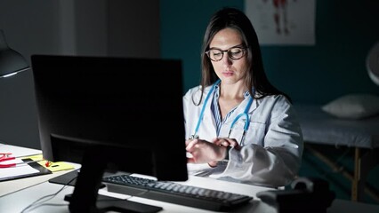 Young beautiful hispanic woman doctor using computer looking clock at clinic