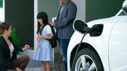Progressive parents with electric vehicle and home charging station. Happy family with daughter giving each other high fives before leave for school. Alternative future transportation concept of EVs