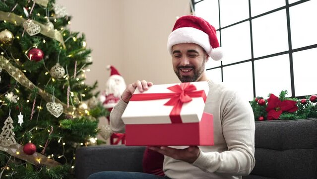 Young hispanic man unpacking gift with unhappy expression sitting on sofa by christmas tree at home