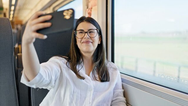 Young beautiful hispanic woman smiling taking a selfie picture with the smartphone sitting at train station