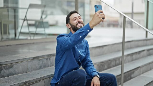 Young hispanic man business worker sitting on stairs make selfie by smartphone at street