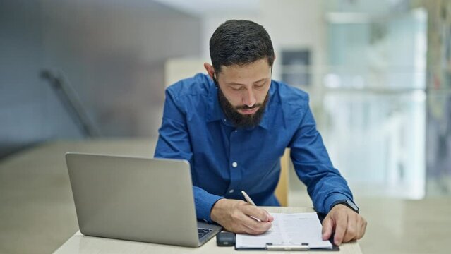 Young hispanic man business worker using laptop and earphones taking notes at the office