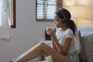 Young African woman drinking coffee with earphones sitting on sofa at home.