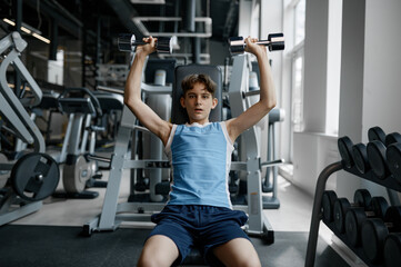 Fototapeta na wymiar Serious strong teenager boy training with dumbbells while sitting on bench