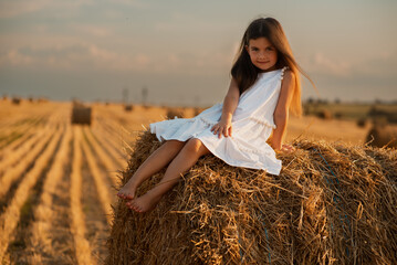 Naklejka na ściany i meble A cute little European-looking girl in a white summer dress sits on a bale of hay