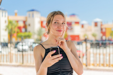 Young blonde woman at outdoors using mobile phone and thinking