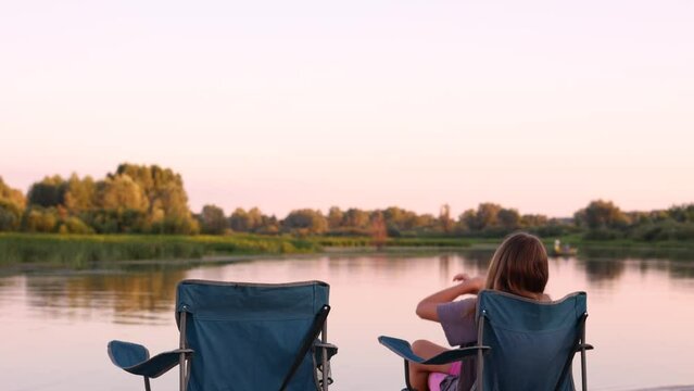 View from back. Girl sits on chair on the river bank enjoying magic red sunset, girl stretching hands