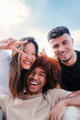 Vertical portrait of group of young multiracial friends hugging and having fun together. Three happy cool buddies smiling looking at camera. Diverse student teenage people laughing bonding at weekend