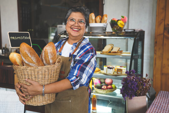 Portrait of a smiling senior Asian barista, hands holding bread basket with promotion sign welcoming the customer in a cozy coffee shop.  Small business owner, family manage.