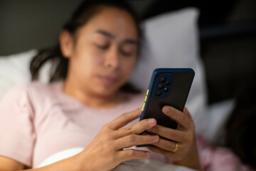 Young Asian woman lying on white bed and playing smartphone at night. Woman in bed with phone. Health and social concept.