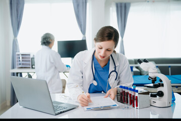Fototapeta premium Confident young Caucasian female doctor in white medical uniform sit at desk working on computer. Smiling use laptop write in medical journal