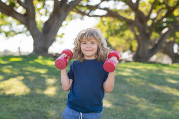 Kids sport training in summer park outdoor. Cute child boy pumping up arm muscles with dumbbell. Fitness kids with dumbbells.